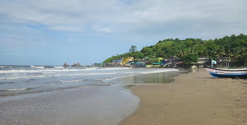 Scenic view of a sandy beach in Goa with gentle waves, colorful shacks along the shoreline, and fishing boats by the coast under a partly cloudy sky.